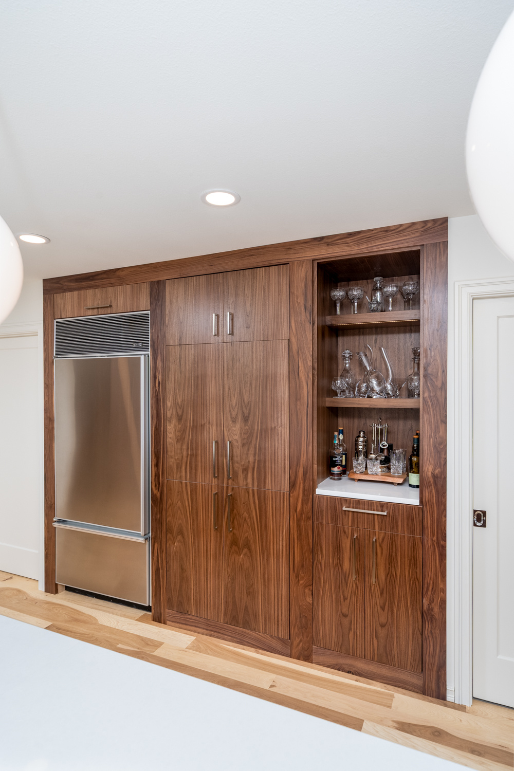 Sleek kitchen design with wood-paneled cabinets, a hidden refrigerator, and a built-in bar with glassware, designed for a whole home remodel in Portland, OR by Creekstone Design + Remodel